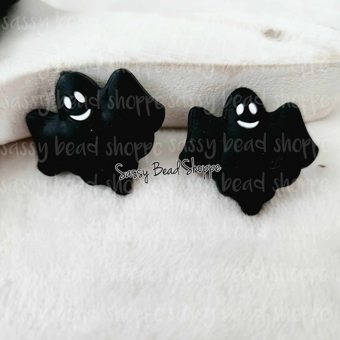 May include: Two black ghost shaped charms with white smiling faces. The charms are on a white fuzzy surface.