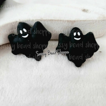 May include: Two black ghost shaped charms with white smiling faces. The charms are on a white fuzzy surface.